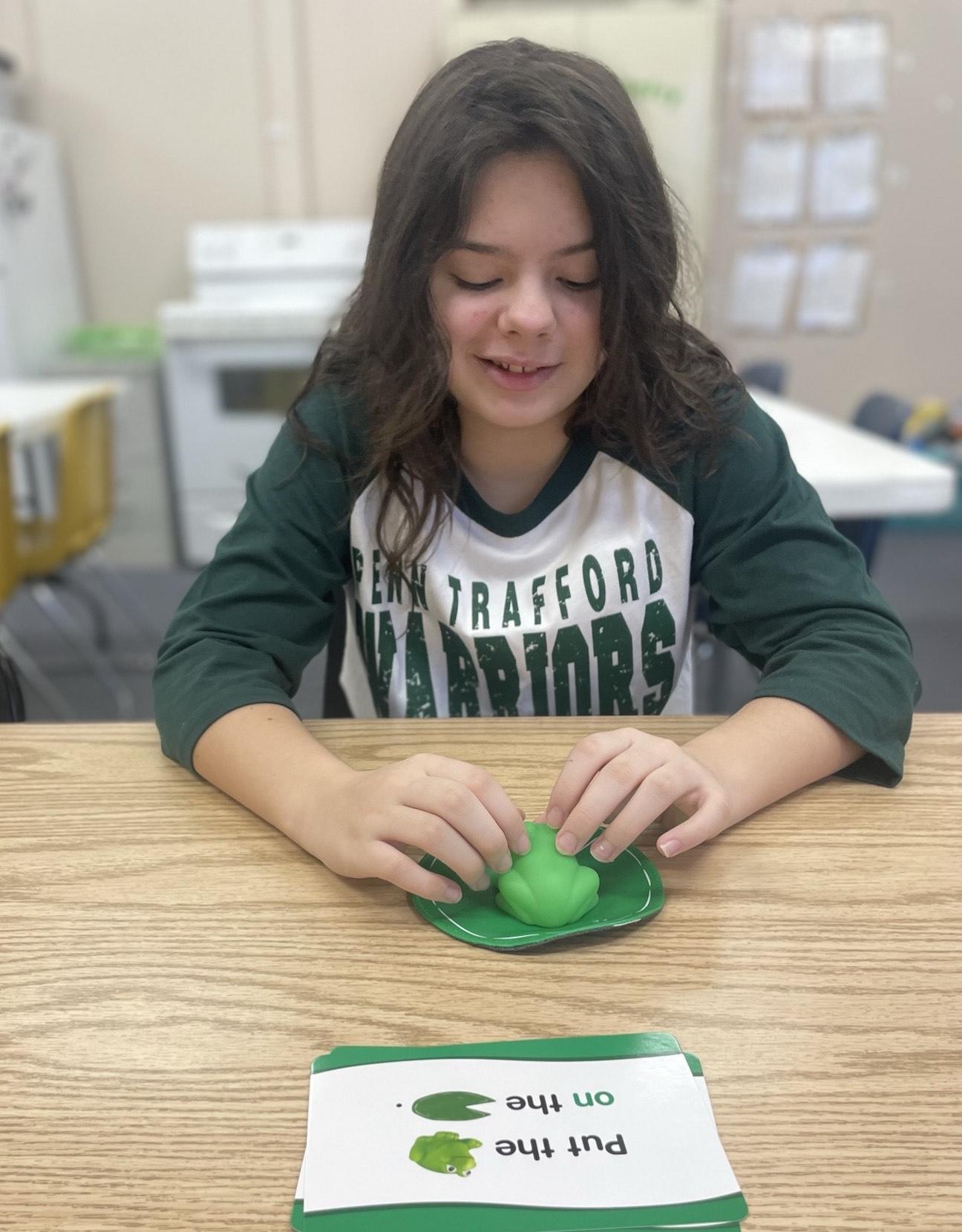 Gianna Acker works on learning new spatial/positional terms such as “on, under, beside, to the left/right” by moving the frog and other objects to different locations